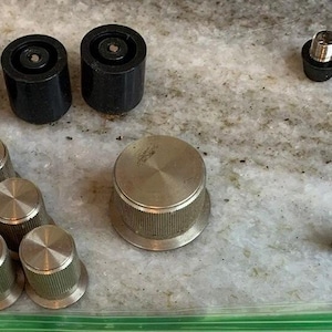 Puede incluir: Colección de varios componentes electrónicos negros y plateados, incluyendo perillas, piezas cilíndricas y un fusible. Los componentes están dispuestos sobre una superficie clara, probablemente para reparación o montaje.