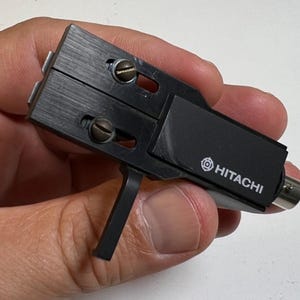 Può includere: Una testina per giradischi Hitachi nera con corpo rettangolare in metallo spazzolato, due viti a taglio visibili per la regolazione e un braccio dello stilo rivolto verso il basso. Il marchio Hitachi è stampato in bianco sul lato.