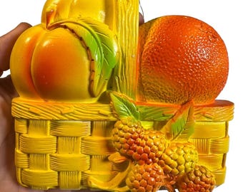 May include: A yellow wall plaque shaped like a woven basket filled with fruit. The basket contains a peach with a green leaf, a textured orange, and a cluster of orange berries with green leaves. The piece features a vertical handle at the top.