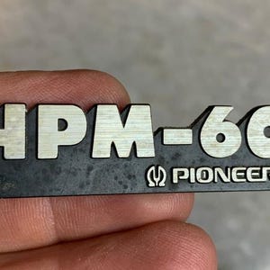 Puede incluir: Un emblema rectangular negro y plateado Pioneer HPM-60. El texto "HPM-60" está en letras plateadas, con "PIONEER" debajo. El emblema está ligeramente desgastado.