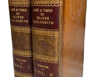 May include: Two antique leather-bound books titled "Life & Times of Oliver Goldsmith" by Forster, Volumes I and II. The spines are burgundy with gold lettering and decorative patterns. The books are from 1854.