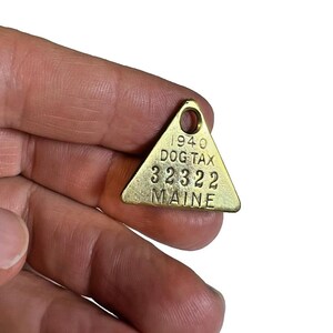 May include: A vintage brass dog tag from 1940, triangular in shape, with the text "DOG TAX 32322 MAINE." The tag has a small hole at the top for attachment. The tag is held in a hand.