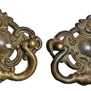 May include: Two antique brass drawer pulls with elaborate scrollwork. Each pull has a central oval plate, a curved handle, and floral detailing. The brass has a warm, aged patina, giving a vintage appearance. Suitable for furniture restoration.