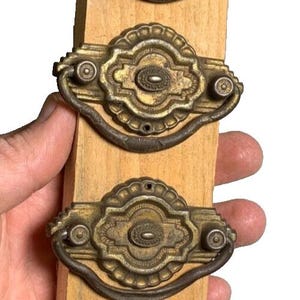 Puede incluir: Seis tiradores de puerta de armario de latón antiguo con diseños ornamentados y un asa de bucle. Los tiradores se muestran en una tabla de madera.