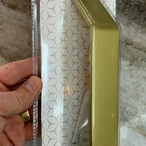 Puede incluir: Una manilla de puerta Charleston de 25,4 cm en oro cepillado para puertas de granero. La manilla está empaquetada en una bolsa de plástico transparente con un fondo estampado blanco y gris. El paquete incluye el texto "National Hardware" y "N700-105".