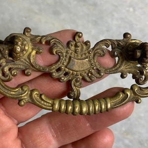May include: Ornate brass drawer pull with a decorative scroll design. The pull has a center section with a raised, ornate design.