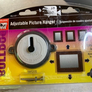 以下が含まれることがあります： 調節可能な絵画ハンガーのパッケージ。ブルドッグのロゴ、"Adjustable Picture Hanger"というテキスト、そして"Push to Rotate"というテキストが書かれたハンガーのクローズアップが特徴です。