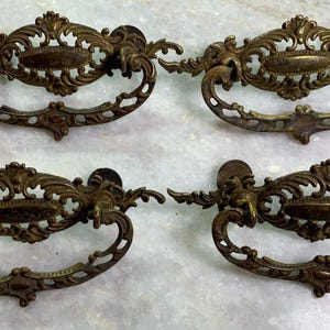 Puede incluir: Cuatro tiradores de cajón de latón antiguo con diseños ornamentados. Cada tirador tiene una forma ovalada decorativa en el centro y una intrincada decoración de volutas.
