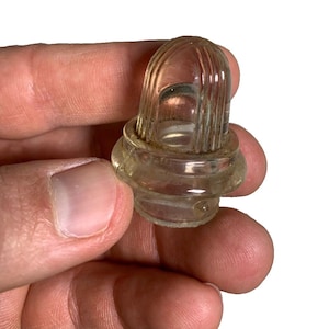 Puede incluir: Un tapón de botella de vidrio transparente con una parte superior acanalada en forma de cúpula. El tapón tiene una base redondeada y está sujeto en la mano de una persona. El vidrio tiene un aspecto ligeramente envejecido.