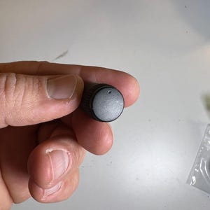 Puede incluir: Un pequeño mando de control negro con un borde texturizado y una parte superior lisa y plana. El mando tiene un pequeño agujero en el centro. El producto es probablemente un componente electrónico o parte de un dispositivo más grande.