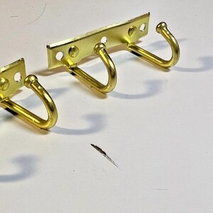 May include: Two brass-colored metal coat racks with three hooks each, and a small pile of screws. The racks have a rectangular base with three holes for mounting. The hooks are curved and rounded.