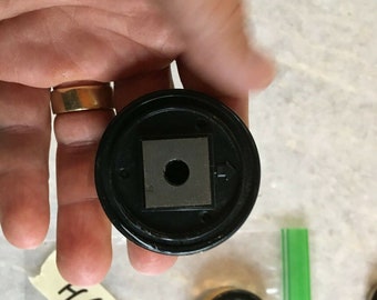 May include: Close-up of a black plastic round object with a square hole in the center. The object has a small black circle in the center of the square hole. The object is being held in a hand.