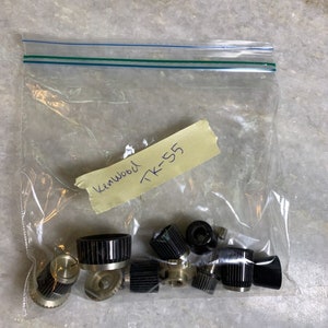 Puede incluir: Una bolsa de plástico transparente que contiene varias perillas y diales negros y plateados. Una cinta adhesiva con el texto escrito a mano "Kenwood TK-55" está adherida. Las perillas parecen ser componentes de equipos electrónicos.