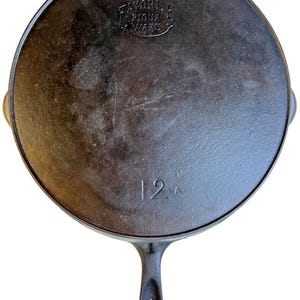 May include: A black cast iron skillet with a long handle and two small side handles. The skillet has the text "FAVORITE PIQUA, O. WARE" and the number "12 A" embossed on it. The skillet is round and has a well-worn appearance.