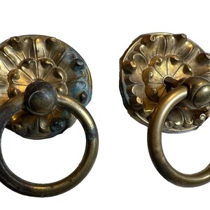 Puede incluir: Dos tiradores de puerta antiguos de latón. Cada tirador presenta un tirador de anillo circular y una placa trasera ornamental con diseño floral. El latón muestra un aspecto envejecido con rastros de verdín.