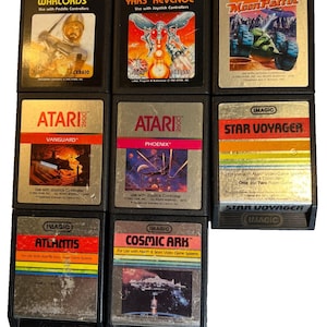 Può includere: Una collezione di cartucce per videogiochi Atari 2600 vintage. I titoli includono "Warlords", "Yars' Revenge", "Moon Patrol", "Vanguard", "Phoenix", "Star Voyager", "Atlantis" e "Cosmic Ark". Le cartucce presentano etichette colorate e custodie in plastica nera.