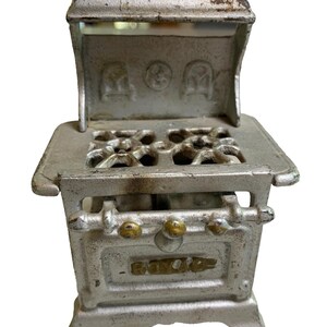May include: A vintage cast iron toy stove with a white painted finish. The stove has a decorative oven door with the word "ROYAL" embossed on it. The stove has a top with four burner openings and a removable hood.
