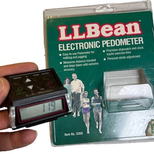 May include: A black LLBean electronic pedometer with a digital display showing "119". The pedometer is held in front of its original packaging, which is green and white and features the LLBean logo and product information.