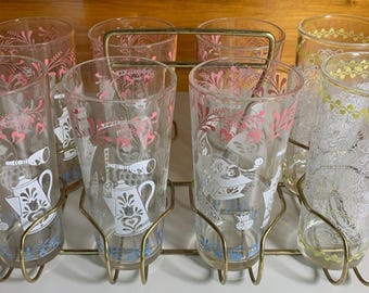 May include: Set of eight clear glass tumblers with a gold metal rack. The glasses have a pink and white floral design with a blue and white teapot motif.