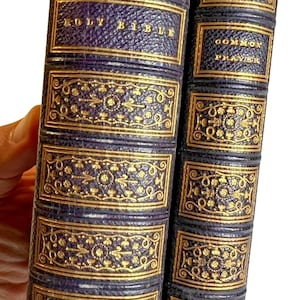 Puede incluir: Dos libros antiguos con cubiertas de cuero morado oscuro y lomos decorativos con relieve dorado. El libro más grande se titula HOLY BIBLE y el más pequeño COMMON PRAYER. Ambos lomos presentan intrincados patrones florales y geométricos.