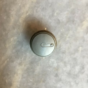 Puede incluir: Un pomo plateado con un pequeño botón redondo gris en el centro.