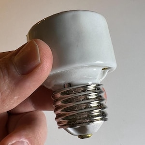 Op de afbeelding: Een witte keramische lampfitting met een zilveren metalen schroefbasis. De fitting wordt in een hand gehouden tegen een neutrale achtergrond. De fitting is ontworpen om een standaard gloeilamp te bevatten.