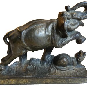 May include: Bronze sculpture of an elephant leaping over a small animal. The elephant has its trunk raised and is in mid-air, with detailed texture. The base is rectangular and the overall colour is a dark bronze.