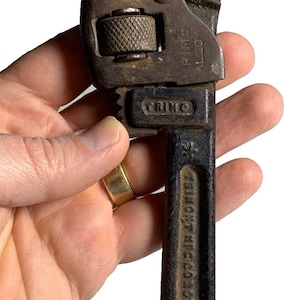 Puede incluir: Una llave inglesa negra y oxidada con el texto "THE TRIMO CO. CHICAGO, ILL. USA" y "ALLOY RIMC" en el mango.