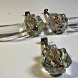 May include: A collection of clear glass cabinet knobs and pulls. The knobs have a faceted, flower-like design with a silver-colored screw. The pulls are curved and rectangular. The hardware is likely for furniture.