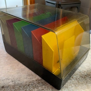 Peut inclure: Une boîte de rangement en plastique transparent avec une base noire et des compartiments de couleurs arc-en-ciel. Les compartiments sont jaunes, rouges, bleus, verts et violets. La boîte est étiquetée "E-Z-Sort".