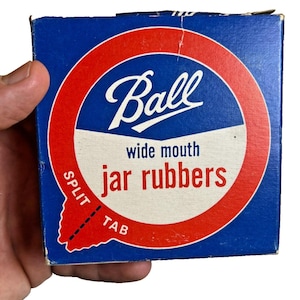 Op de afbeelding: Een blauwe doos met Ball wide mouth potrubbers. De doos heeft een rood en wit cirkelvormig ontwerp met het Ball-logo in witte letters. De woorden "wide mouth" en "jar rubbers" zijn ook zichtbaar. De doos heeft een split tab.