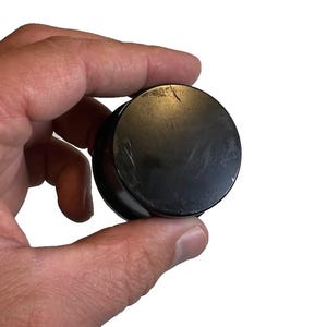 Puede incluir: Un objeto negro y redondo con una parte superior plana y un borde ligeramente elevado. El objeto se sostiene en una mano sobre un fondo blanco. La superficie del objeto tiene algunos arañazos visibles.
