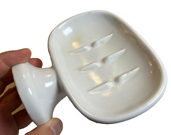 May include: A white ceramic soap dish with a curved handle. The dish has a rectangular shape with four raised, angled ridges for drainage. The glossy finish reflects light, highlighting the smooth surface of the bathroom accessory.