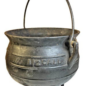 May include: A black cast iron cauldron with a rounded body, three short legs, and a thin metal handle. The exterior features embossed text that reads 1/2 GALL, indicating its capacity. The surface has a textured, dark, and weathered appearance.