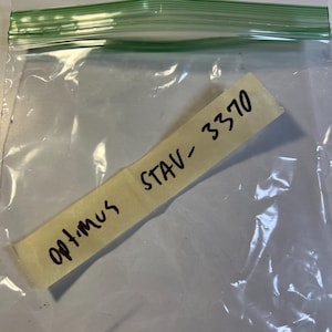 Puede incluir: Una bolsa de plástico transparente con una parte superior sellada verde. Dentro de la bolsa hay una etiqueta amarilla con el texto manuscrito "Optimus STAV-3370" en tinta negra. La etiqueta está ligeramente arrugada.