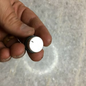 Puede incluir: Un pomo redondo blanco con una pequeña línea negra en él. El pomo está unido a una base de metal plateado.