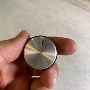 Puede incluir: Un pomo plateado con un anillo negro alrededor del borde. El pomo tiene una pequeña línea negra en la parte superior.