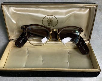 May include: A pair of gold-rimmed glasses with brown frames and green accents in a black and gold case. The case has a satin lining and the glasses have the word "Zenith" written on the side.