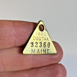 May include: A vintage brass dog tag from 1940, triangular in shape. It has a hole for attachment and the text "DOG TAX", "32360", and "MAINE". The tag is held in a person's hand.