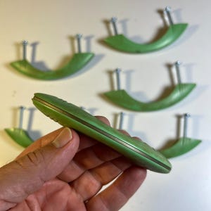 May include: A set of five sage green, crescent-shaped drawer pulls. Each handle features a smooth, curved design with a metallic silver-toned edge and two protruding metal mounting screws on the back for installation purposes.