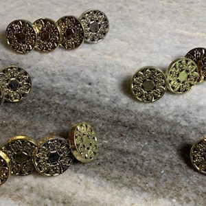 Peut inclure: Un ensemble de 10 boutons décoratifs en métal doré avec des motifs complexes. Les boutons sont disposés en trois groupes, chaque groupe présentant un motif différent.