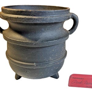 May include: A black cast iron cauldron with two handles and three small feet. The cauldron has a textured surface and a rounded shape. A pink eraser with the text "Pink Pearl" is next to the cauldron.