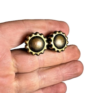 Pode incluir: Dois brincos de botão antigos dourados. Cada brinco apresenta um design circular central, ligeiramente abobadado, rodeado por uma borda ondulada. Os brincos são segurados na mão de uma pessoa contra um fundo branco.