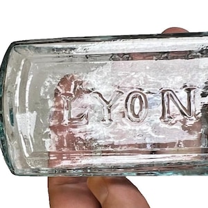May include: A clear, rectangular glass bottle with the embossed text "LYON'S". The bottle has a rounded neck and a flared, ridged stopper. The bottle is held in a person's hand.