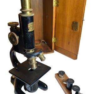 May include: An antique microscope with a black body and brass accents, accompanied by a wooden case and a wooden holder with lenses. The microscope has the text "GRA APSO" and "BAUSCH & LOMB" printed on it.
