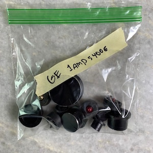 Puede incluir: Una bolsa de plástico que contiene un conjunto de perillas de estufa negras y rojas. La bolsa está etiquetada "GE 1AMP 5400E".