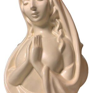 May include: A white ceramic sculpture of a woman with her hands clasped in prayer. She is wearing a long, flowing robe and has a serene expression on her face.