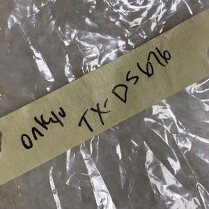 Puede incluir: Una etiqueta de papel marrón con el texto "олкуч TX-DS676" escrito en marcador negro.