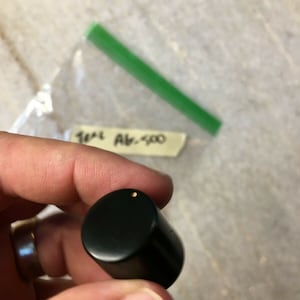 Puede incluir: Una perilla negra con un pequeño punto dorado en el centro.