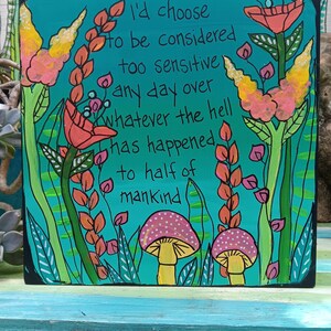 May include: A square, hand-painted wooden sign with a teal background and black border. The sign features colorful floral illustrations and the text "I'd choose to be considered too sensitive any day over whatever the hell has happened to half of mankind."